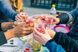 Several people raising plastic cups filled with sparkling drinks and strawberries for a toast at an outdoor gathering.