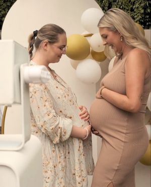 Two smiling pregnant women stand facing each other, looking down. They are both holding their baby bumps. The woman on the left wears a light-colored, floral dress and round glasses, her hair in a high braided ponytail. The woman on the right wears a fitted, beige ribbed sleeveless dress. The background features a white round backdrop arch decorated with clusters of white and gold-colored balloons. In the left corner of the photo, a part of a white photo booth is visible in the foreground.