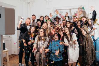 Una foto vivace del team di KRUU che festeggia davanti a una cabina fotografica bianca situata sulla sinistra. I dipendenti ridono e lanciano coriandoli colorati verso la macchina fotografica mentre indossano accessori festosi come occhiali oversize. La scena cattura un senso di comunità energica e gioiosa.