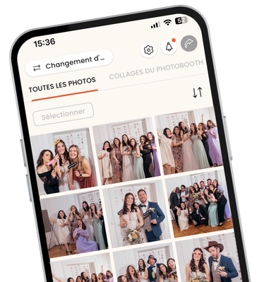 Vue rapprochée et inclinée d'un smartphone affichant l'interface de l'application mobile KRUU. L'écran affiche une galerie de photos numériques intitulée "Alle Bilder" (Toutes les images) remplie de diverses photos de groupe prises lors d'un mariage. Les photos montrent des invités en tenue de soirée qui posent avec des accessoires amusants tels que des verres en forme de cœur et des panneaux "Just Married". Le design de l'application est propre et moderne, avec des icônes de navigation pour les paramètres, les notifications et le partage en haut de l'application.