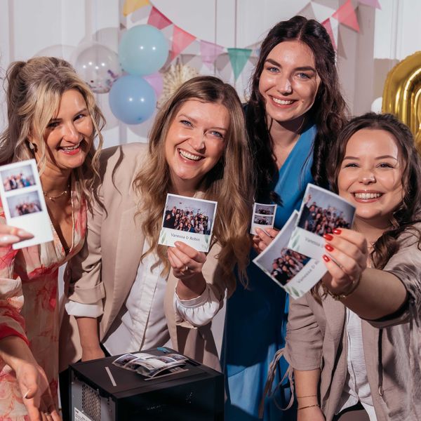 Four smiling women in formal attire are holding up various photo booth prints toward the camera. The prints feature group photos and are personalized with the names "Vanessa & Robin." In the foreground, a photo printer is visible with more photo strips resting on top. The background is festively decorated with light blue and golden foil balloons as well as a colorful pennant banner.