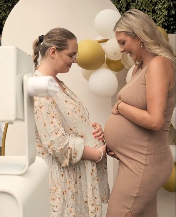 Two smiling pregnant women stand facing each other, looking down. They are both holding their baby bumps. The woman on the left wears a light-colored, floral dress and round glasses, her hair in a high braided ponytail. The woman on the right wears a fitted, beige ribbed sleeveless dress. The background features a white round backdrop arch decorated with clusters of white and gold-colored balloons. In the left corner of the photo, a part of a white photo booth is visible in the foreground.